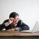 Modern Asian man in jacket and glasses looking at laptop and screaming with mouth wide opened on white background